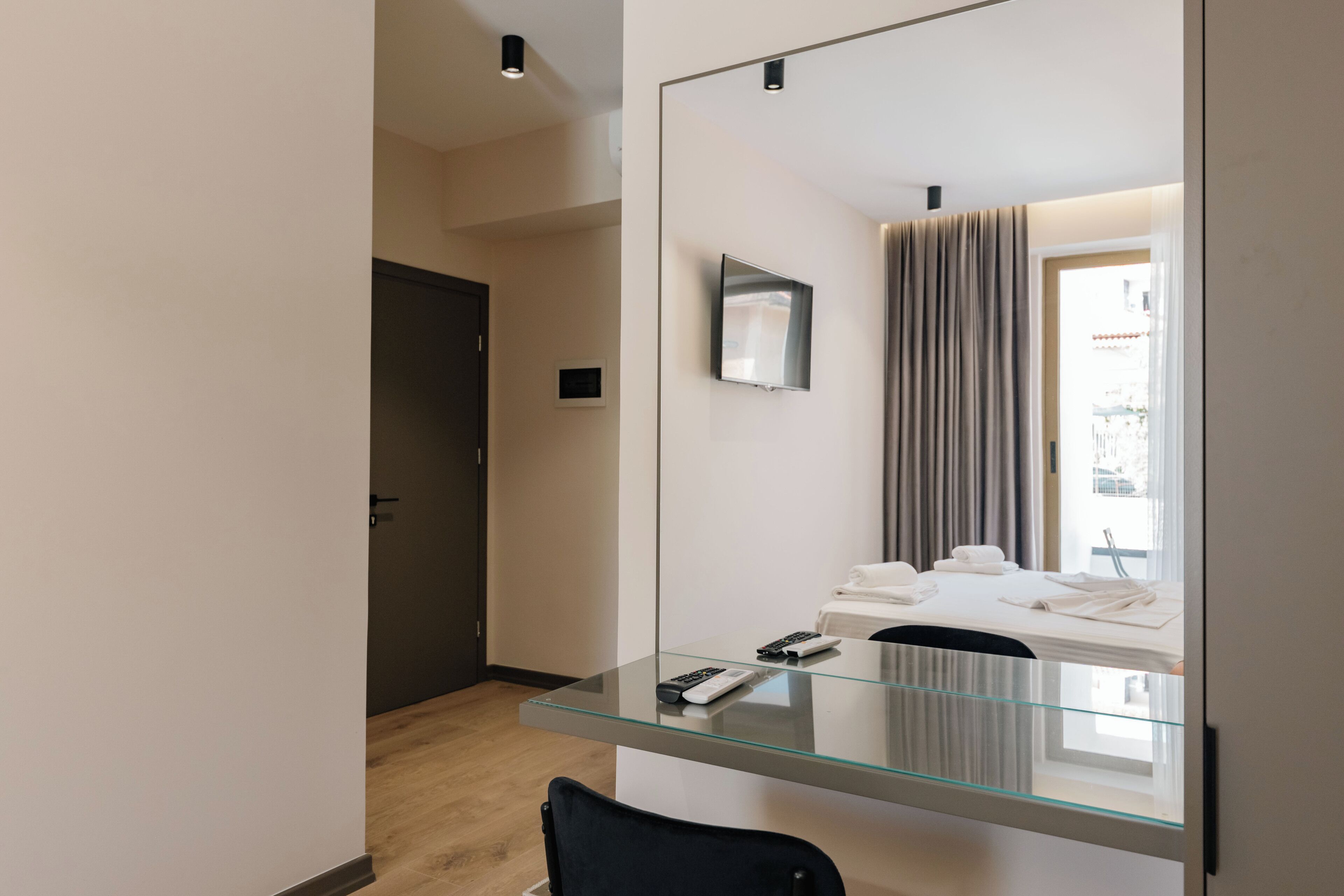 Habitación de hotel moderna con escritorio de vidrio, televisor de pantalla plana, cama con sábanas blancas y un espejo grande que refleja la habitación.
