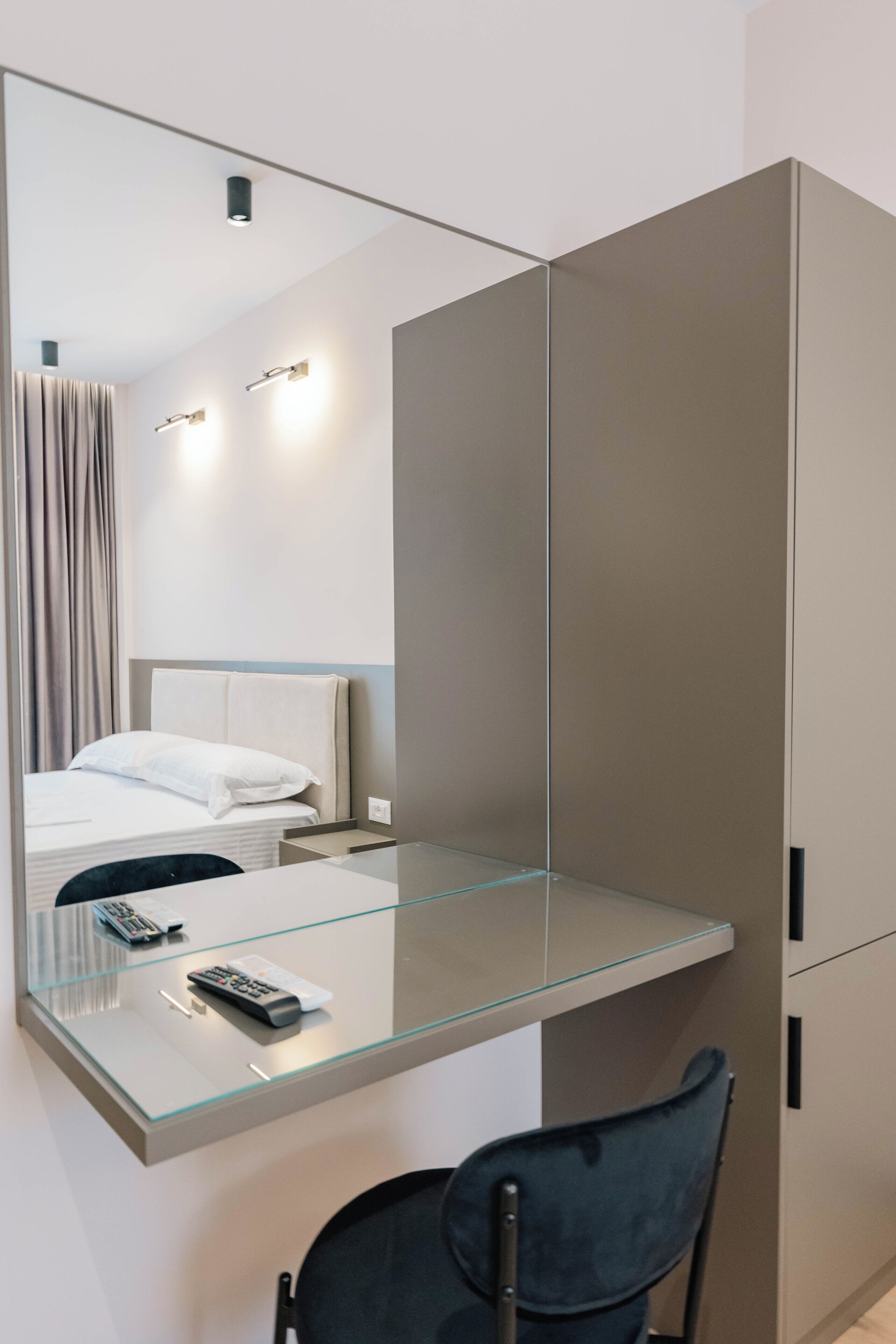A modern hotel room with a glass desk, a chair, a bed with white linens, and a large mirror reflecting the room.