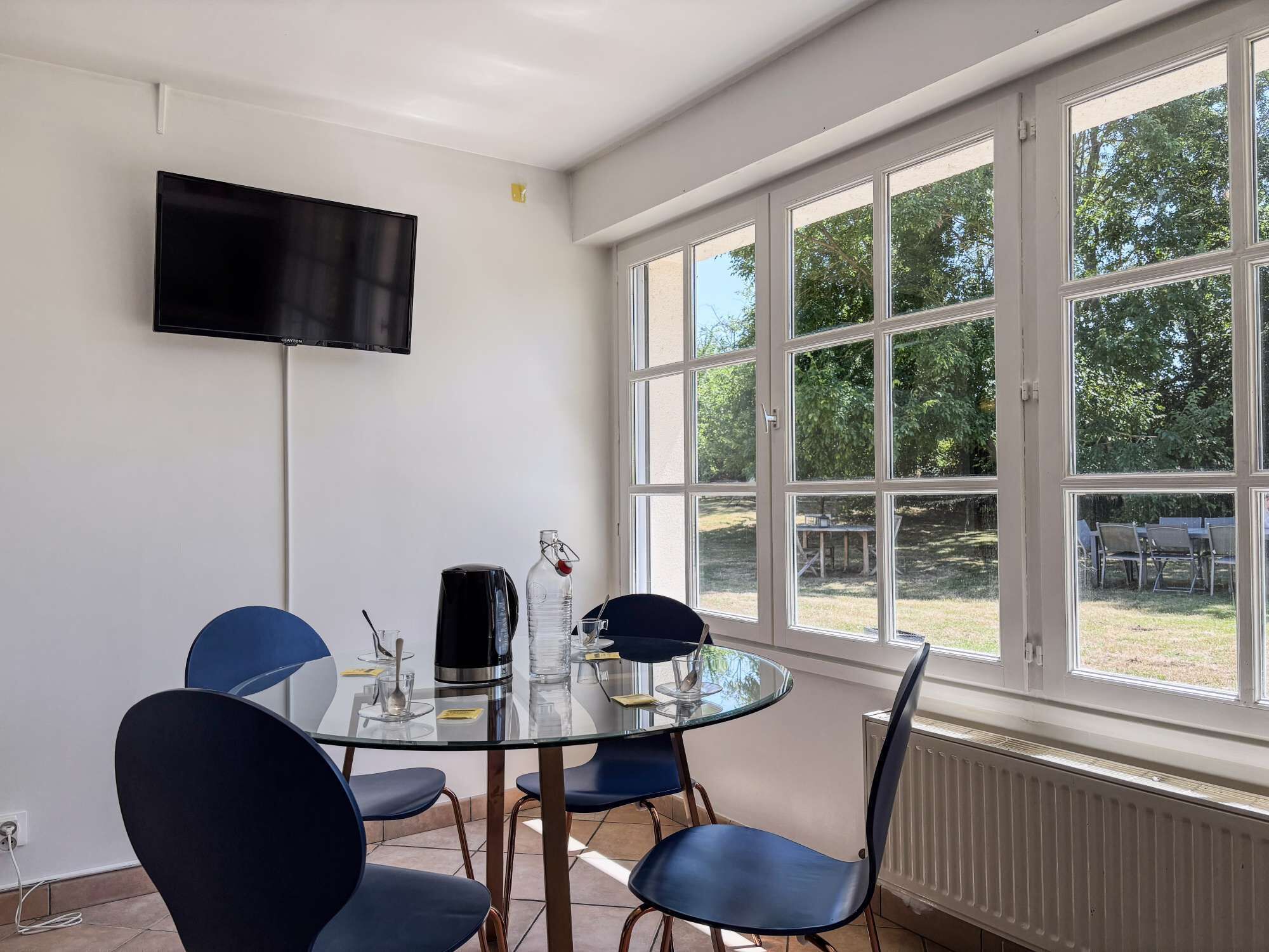 A conference room with a glass table, blue chairs, a black coffee maker, a bottle, and a TV on the wall.