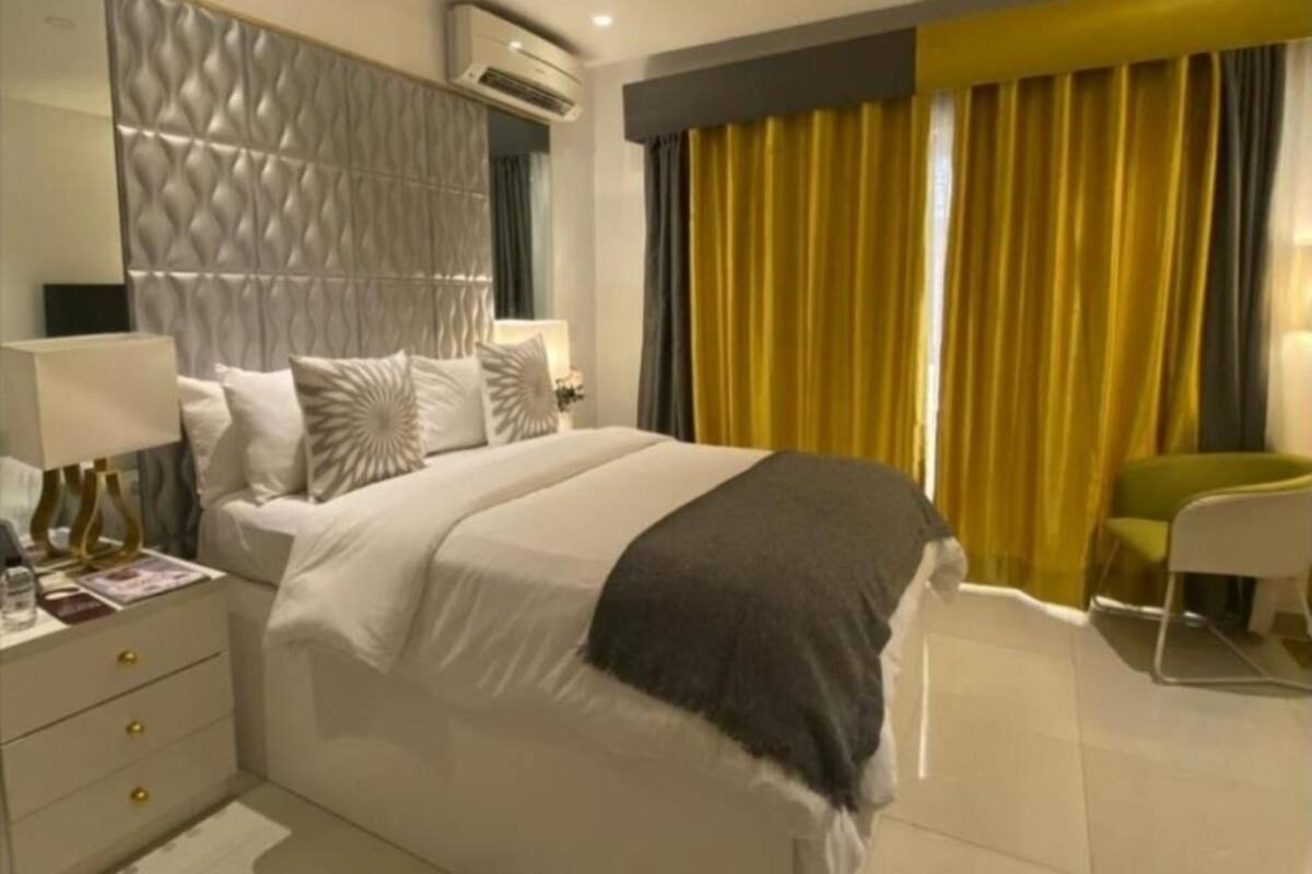 Habitación de hotel moderna con cama, mesitas de noche, una silla y un amplio ventanal con cortinas.