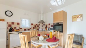 A kitchen with a dining table, chairs, a clock, a microwave, and a stove. - Gîte 'Le Petit Étang' with Shared Pool, Private Terrace and Air Conditioning (Gréoux-les-Bains)