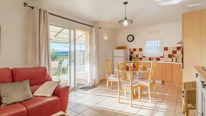 A living room with a red sofa, a dining area with wooden chairs, a kitchen with wooden cabinets, and a view through a sliding glass door. - Gîte 'Le Petit Étang' with Shared Pool, Private Terrace and Air Conditioning (Gréoux-les-Bains)