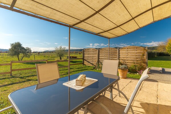 Un área de comedor al aire libre con techo, una mesa y sillas, cerca de una valla de madera, y vistas a un paisaje verde.