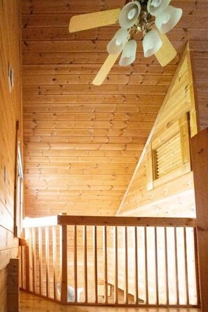 A wooden cabin interior with a sloped ceiling, a ceiling fan, and a wooden railing along the balcony.