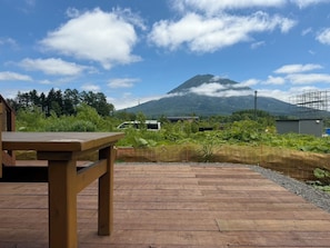 A wooden deck with a table, surrounded by greenery and a mountain in the background. - Powder Hut (Kutchan)