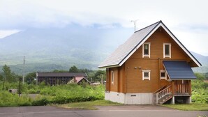 A wooden house with a sloped roof and a small porch, surrounded by greenery and mountains in the background.