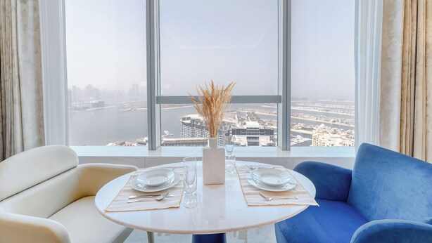 A dining area with a table set for two, a vase with dried plants, and a view of a cityscape through large windows. - PVH - Inviting Studio - Palm Tower (Dubai)