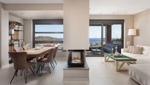A modern living room with a dining area, large windows offering a view of the sea, and a fireplace. - Villa Nefeli Athenian Riviera (Saronikos)