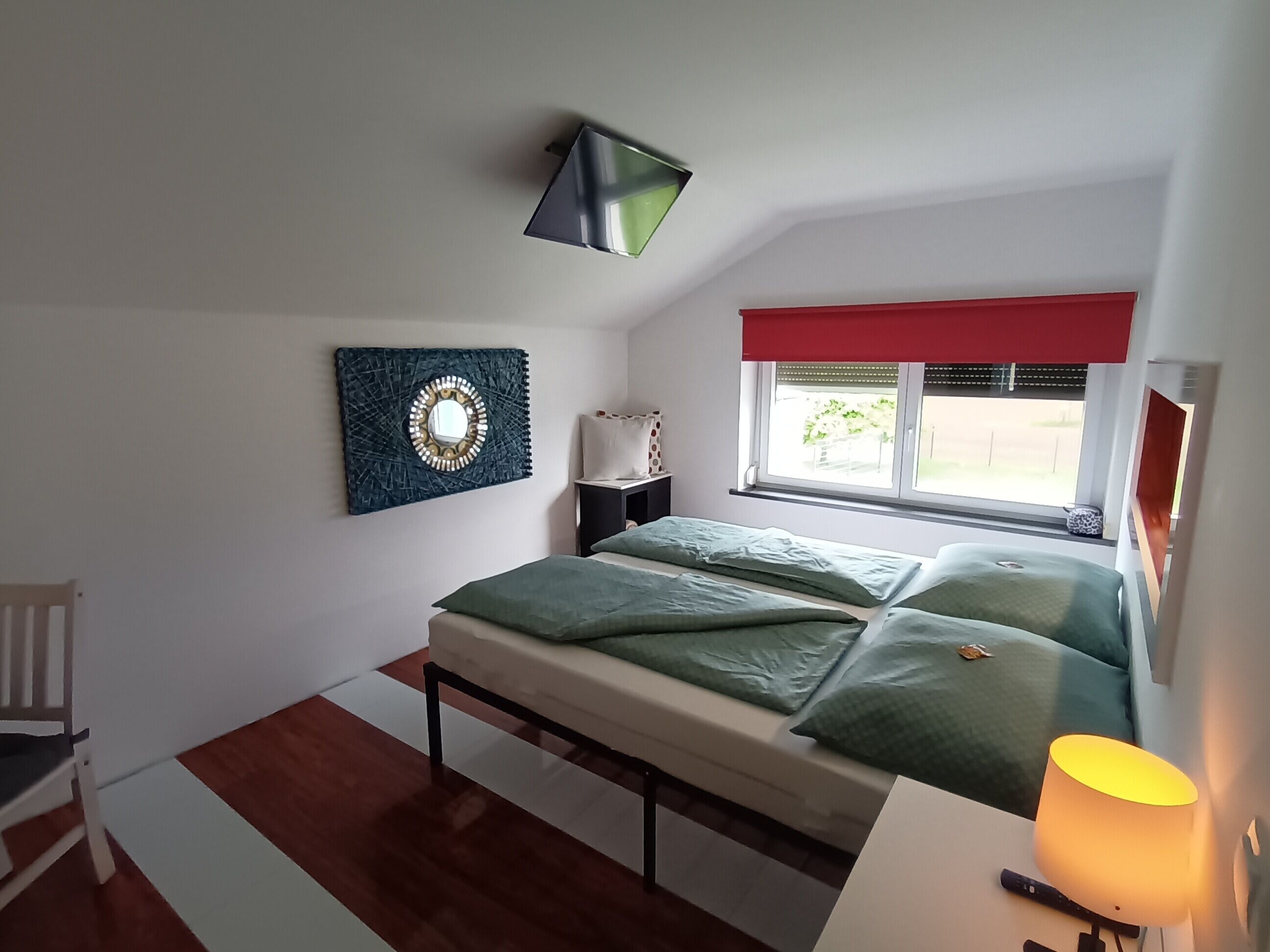 Ein Schlafzimmer mit einem Bett, einem Nachttisch, einem Stuhl, einem Fenster mit roter Vorhang und einer Wandlampe.
