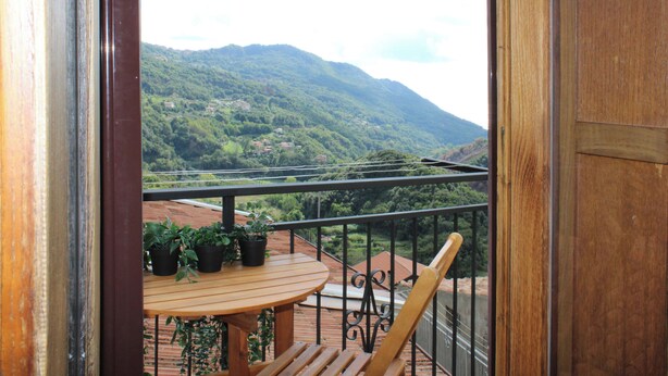 Een balkon met een houten tafel en stoelen, bloempotten en uitzicht op een groene vallei met huizen en een berg.
