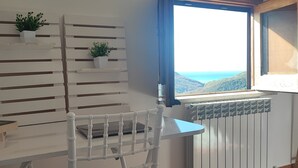 Een kamer met een witte radiator, een raam met uitzicht op zee, een witte stoel en een witte plank met planten.