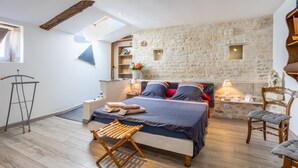 Un dormitorio con pared de piedra, cama con almohadas, una silla, piso de madera y tragaluz.
