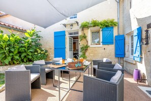 Eine Terrassenausstattung mit Korbmöbeln, einem Tisch mit Korb und Blick auf ein Gebäude mit blauen Fensterläden.