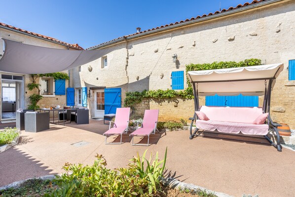Un patio con mobiliario exterior, una zona cubierta y un edificio con contraventanas azules.