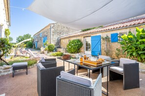 Eine Terrasse im Freien mit Korbmöbeln, einem Tisch mit Korb und Gläsern sowie ein Gebäude mit blauen Fensterläden.