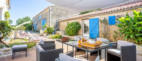 Une terrasse extérieure avec des meubles en osier, une table agrémentée d’un panier, et un bâtiment aux volets bleus.