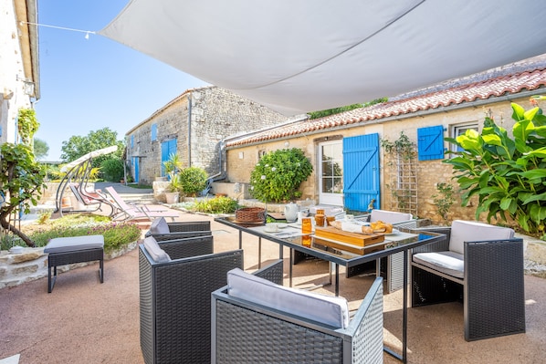 Une terrasse extérieure avec des meubles en osier, une table agrémentée d’un panier, et un bâtiment aux volets bleus.