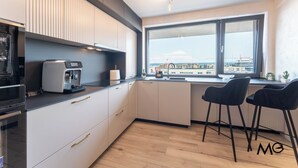 A modern kitchen with a coffee machine, a toaster, and a view of the city through a large window. - MoHost Valcom - Luxury Skyline Penthouse (Brașov)