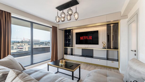 A modern living room with a large TV, comfortable seating, and a view of the cityscape. - MoHost Valcom - Luxury Skyline Penthouse (Brașov)