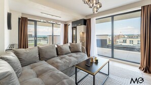 A modern living room with a large sectional sofa, a coffee table, and a view of the city through large windows. - MoHost Valcom - Luxury Skyline Penthouse (Brașov)