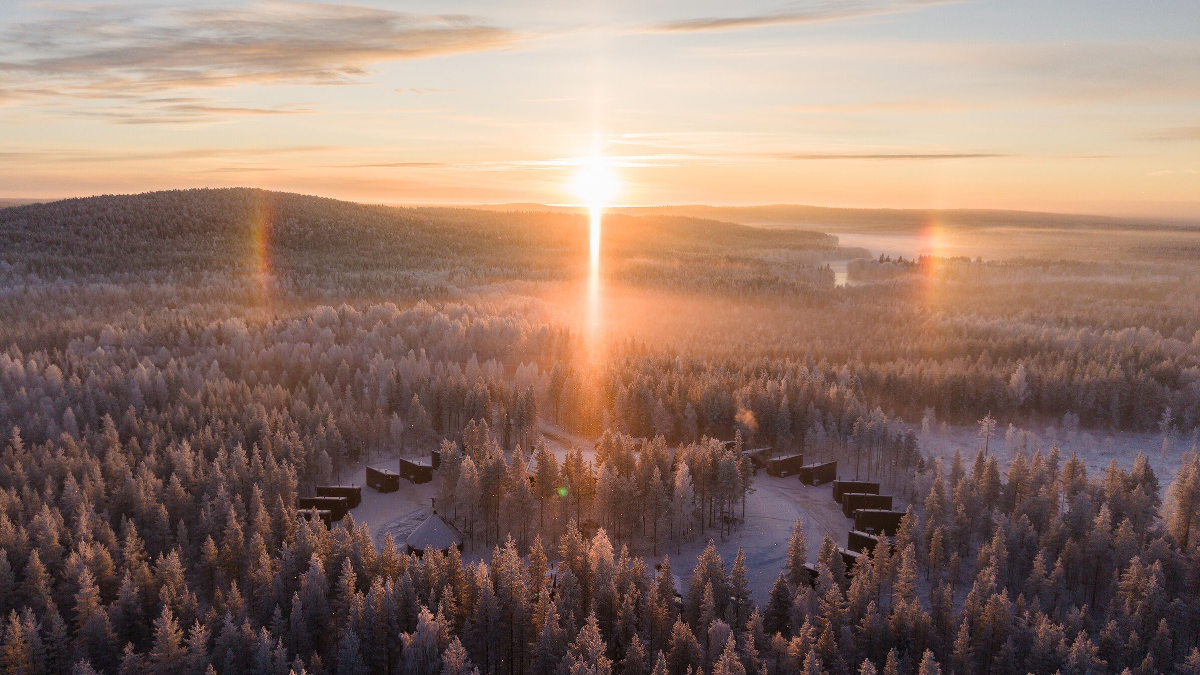 Lumipeitteinen metsä, jossa rakennukset, todennäköisesti mökit, on järjestetty ympyrän muotoon, ja taustalla näkyy auringonnousu.