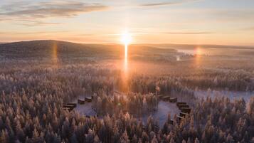 Lumipeitteinen metsä, jossa rakennukset, todennäköisesti mökit, on järjestetty ympyrän muotoon, ja taustalla näkyy auringonnousu.