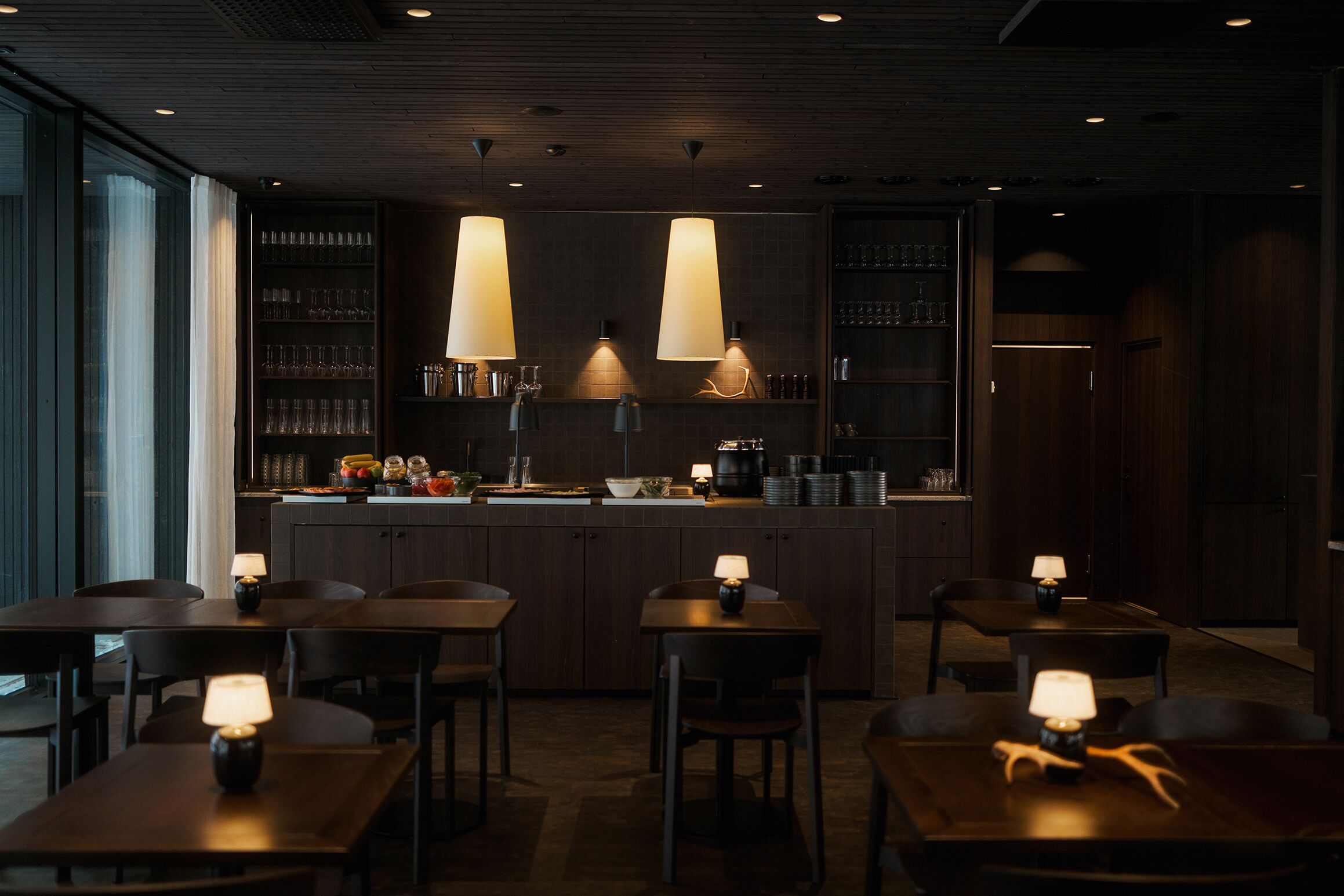 A dimly lit restaurant with wooden tables and chairs, pendant lights, and a bar area with shelves of glasses and bottles.