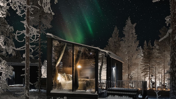 Uma cabana coberta de neve com parede de vidro, uma cama e vista para o céu noturno.