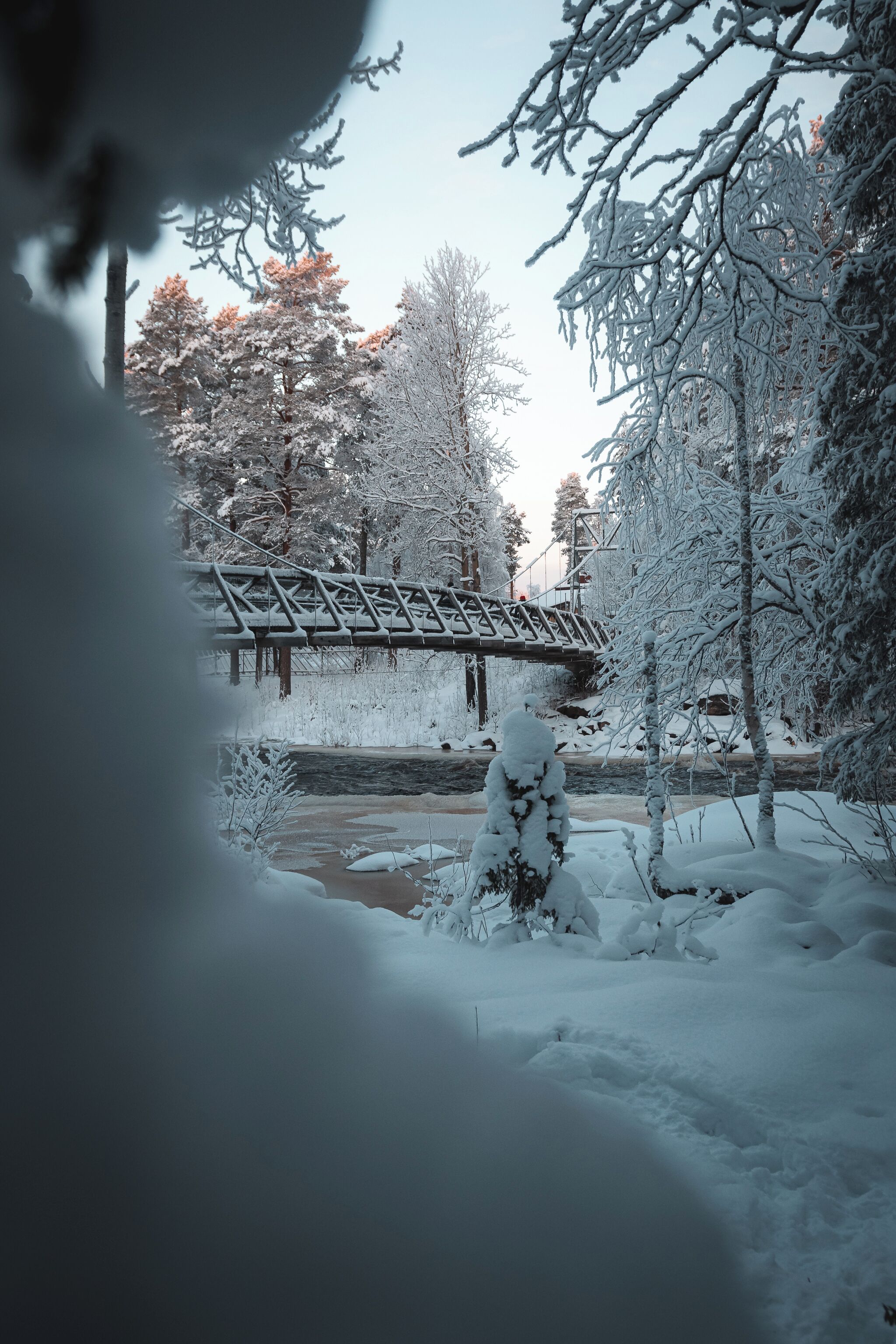 A snow-covered landscape with a bridge, trees, and a building in the background.