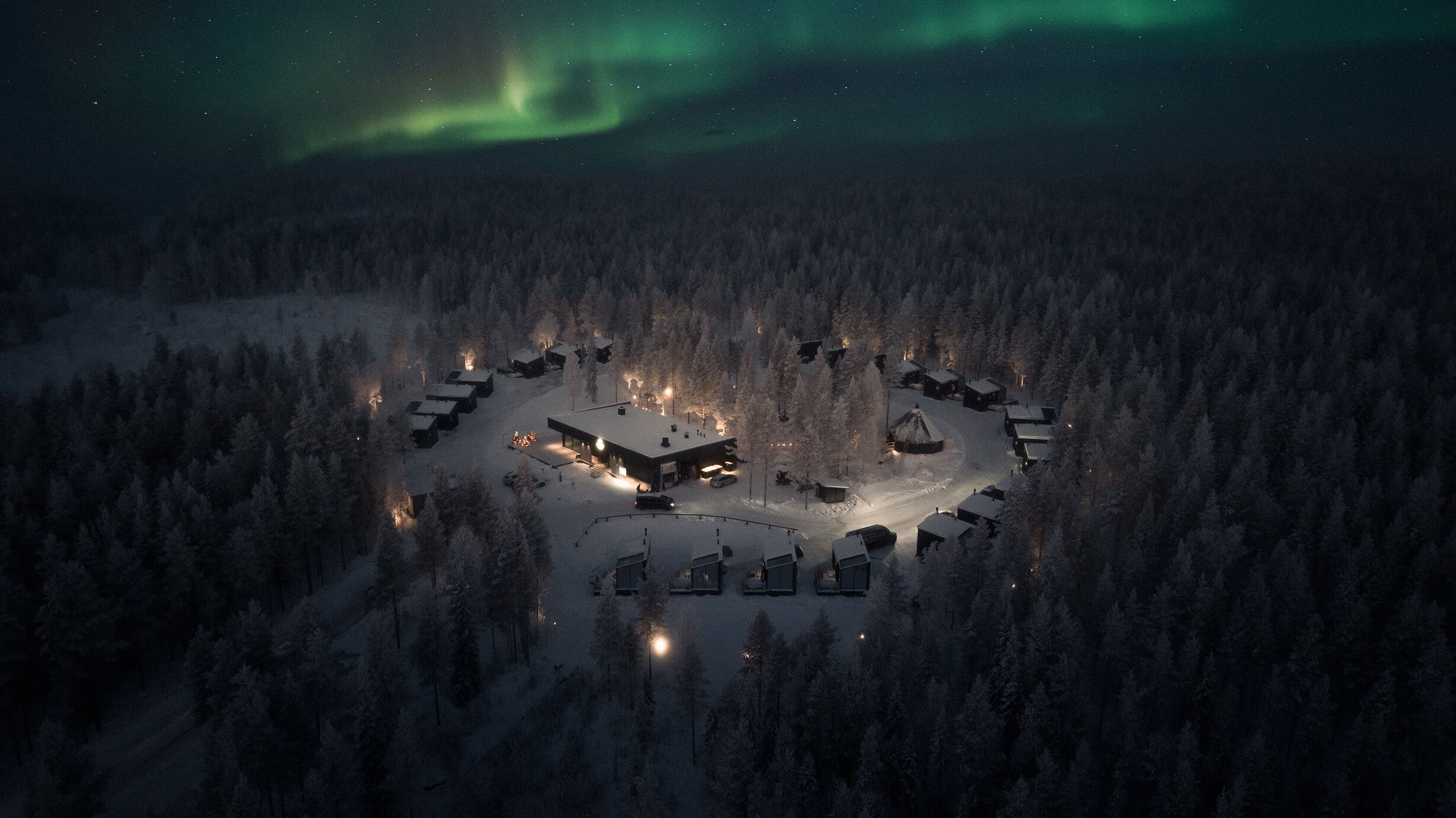A snow-covered forest with a circular arrangement of cabins and a central building, likely a lodge or resort.