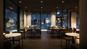 Restaurante com interior moderno, com mesas e cadeiras de madeira, piso de tijolos e grandes janelas que oferecem vista para árvores cobertas de neve.