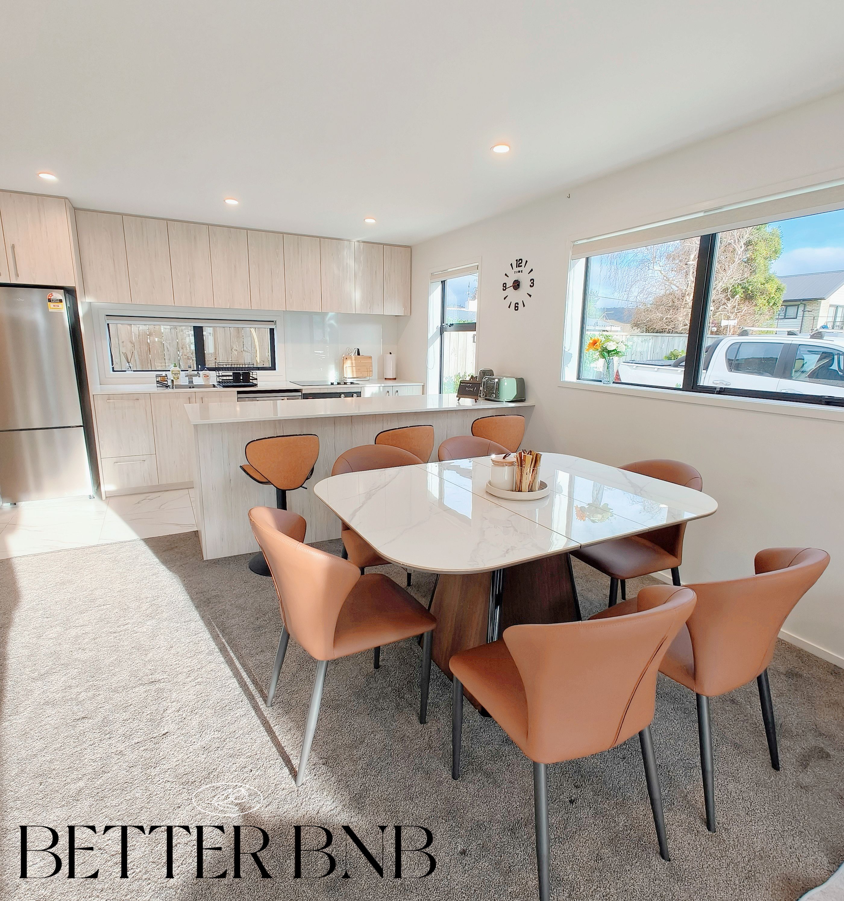 A modern kitchen with a central island, a dining area with a glass table and orange chairs, and a large window with a view of the outside.