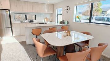 A modern kitchen with a central island, a dining area with a glass table and orange chairs, and a large window with a view of the outside.