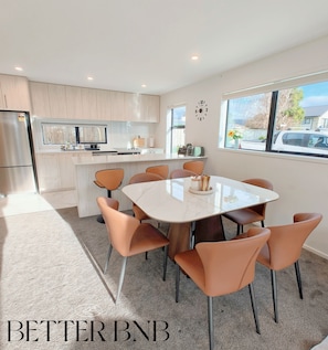A modern kitchen with a central island, a dining area with a glass table and orange chairs, and a large window with a view of the outside. - Modern & New • 3 Beds • 6 Guests (Upper Hutt)
