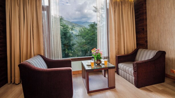 A room with two armchairs, a small table, and a view of a mountainous landscape through a large window with curtains. - TRIBHAV Rohtang Heights Resort (Manali)