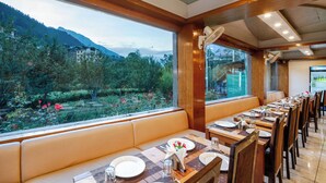 A restaurant with a long table set for dining, a bench seating area, and a view of a mountainous landscape through large windows. - TRIBHAV Rohtang Heights Resort (Manali)