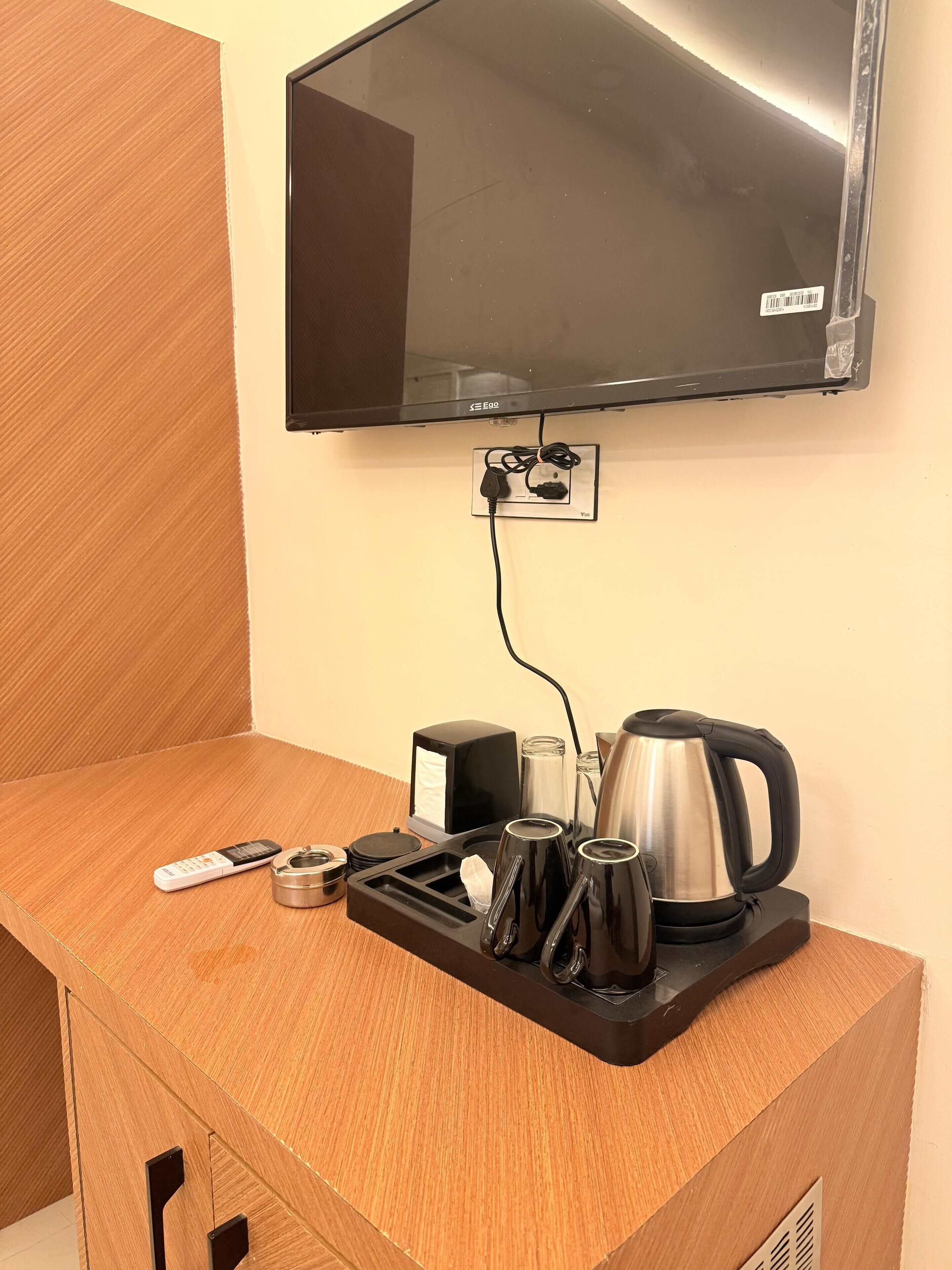 A hotel room with a wooden desk, a television mounted on the wall, a kettle, and a tray with a remote control and a glass.