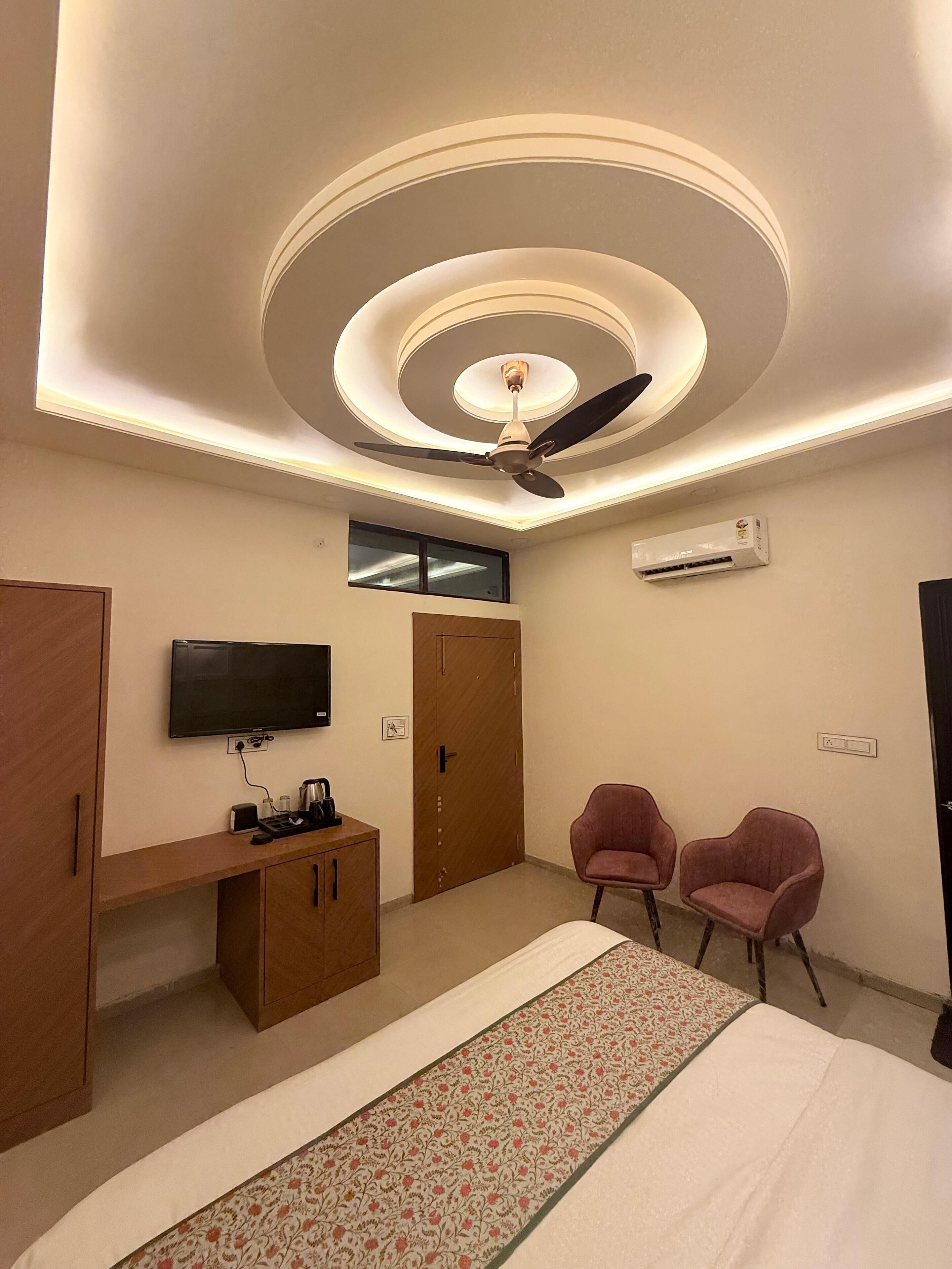 A modern bedroom with a ceiling fan, a flat-screen TV, a wooden wardrobe, two pink chairs, and a bed with a patterned runner.