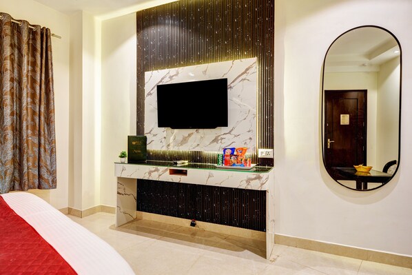 A hotel room with a bed, a TV mounted on a marble wall, a console table with snacks, a mirror, and a door. - Hotel The Krishna (New Delhi)
