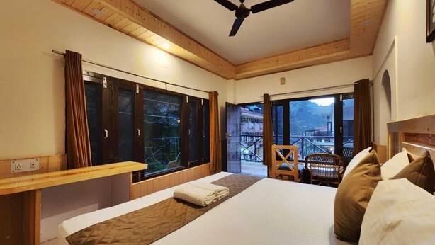 A hotel room with a large bed, a ceiling fan, a window with curtains, and a view of a balcony. - Green Valley Kasol (Kasol)