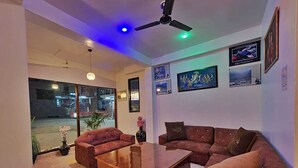 A modern living room with a sectional sofa, a wooden coffee table, and a ceiling fan. There are framed pictures on the wall and a large window providing a view of the street outside. - Green Valley Kasol (Kasol)