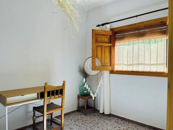 Una habitació amb escriptori de fusta, una cadira, una taula petita amb taulell de vidre, una finestra amb persianes de fusta i un ventilador de sostre.
