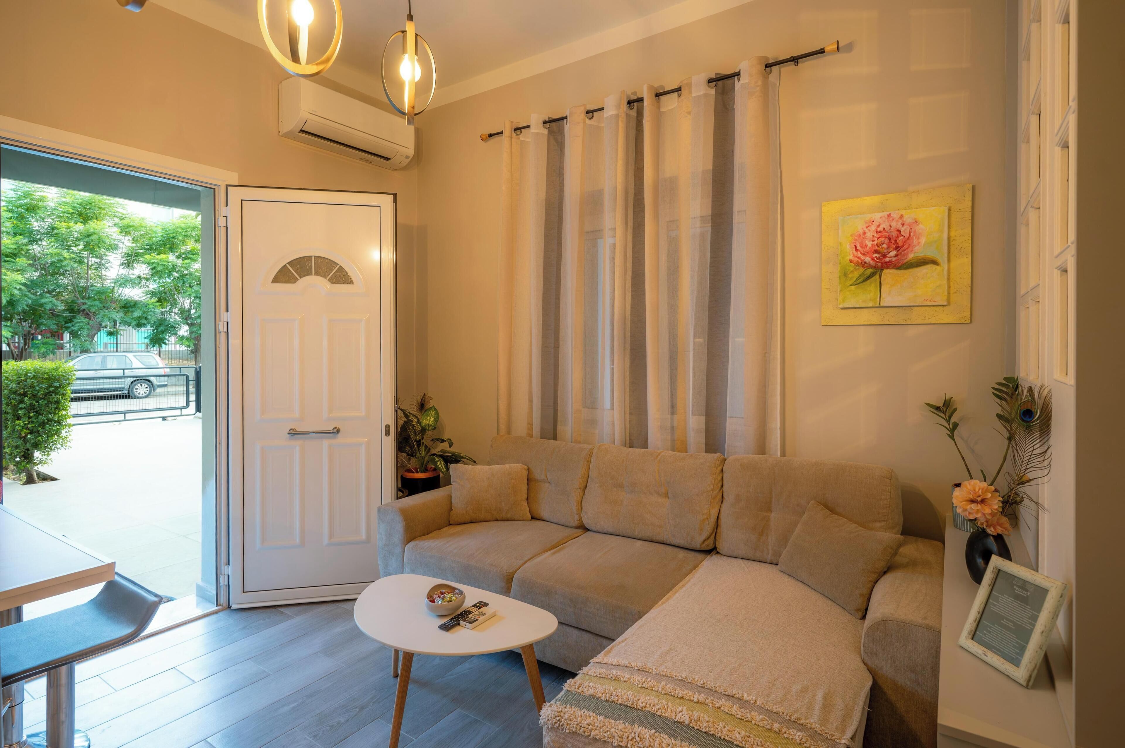 Un salon avec un canapé beige, une petite table blanche et une porte donnant sur l’extérieur. Un tableau représentant une fleur est accroché au mur, et un vase rempli de fleurs est posé sur une table d’appoint.