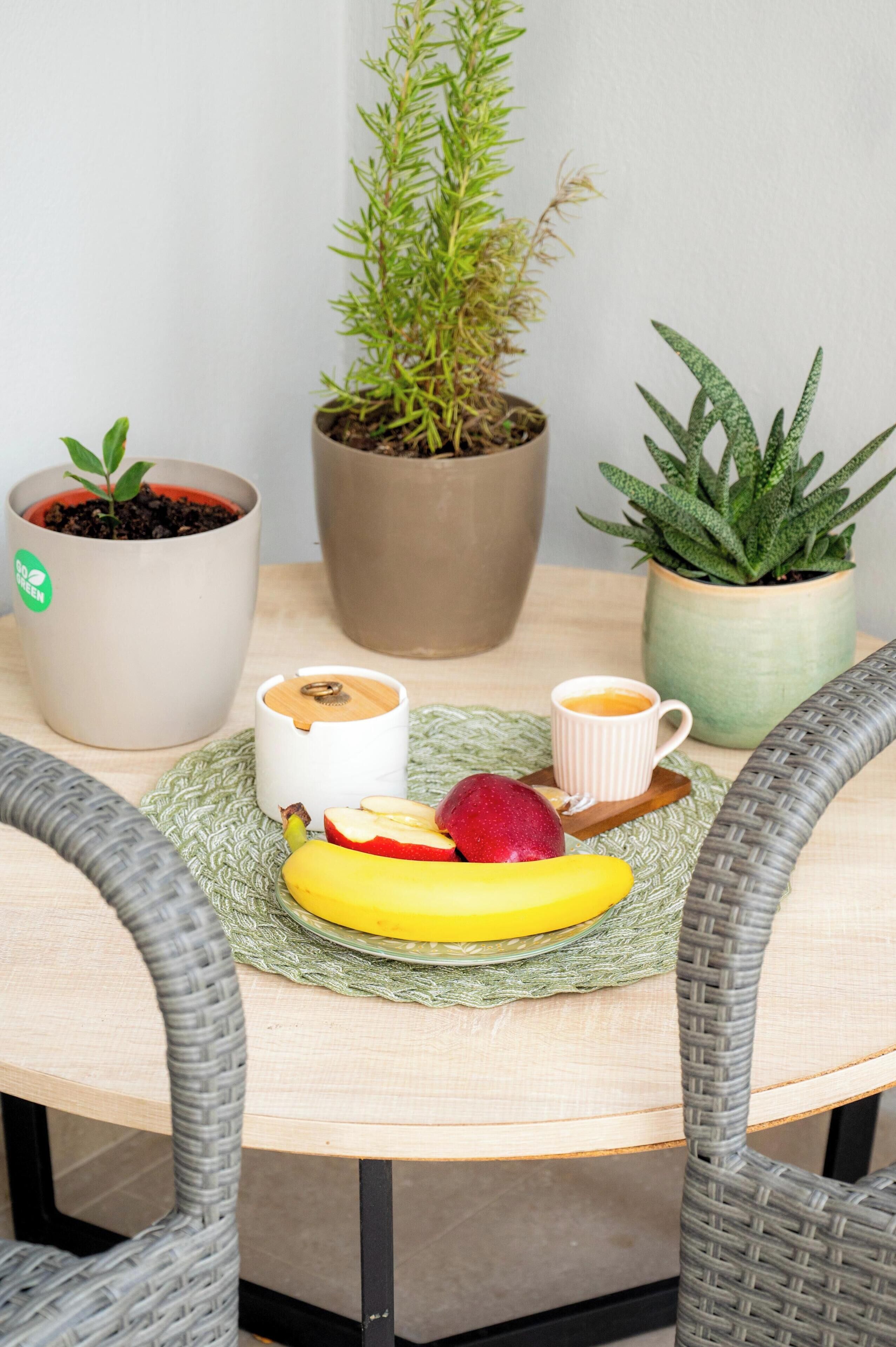 A round table with a woven placemat, two potted plants, a cup of coffee, a banana, and a red apple.