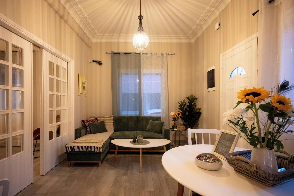 Una sala de estar acogedora con un sofá verde, una mesa de centro blanca y una mesa de comedor con un jarrón de girasoles.