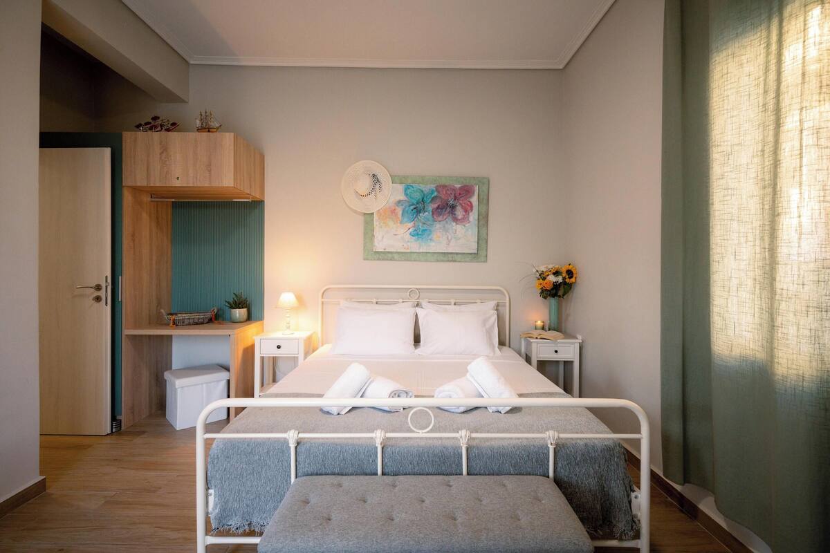 Un dormitorio ordenado con cama, mesitas de noche, un escritorio pequeño y una ventana con cortinas.
