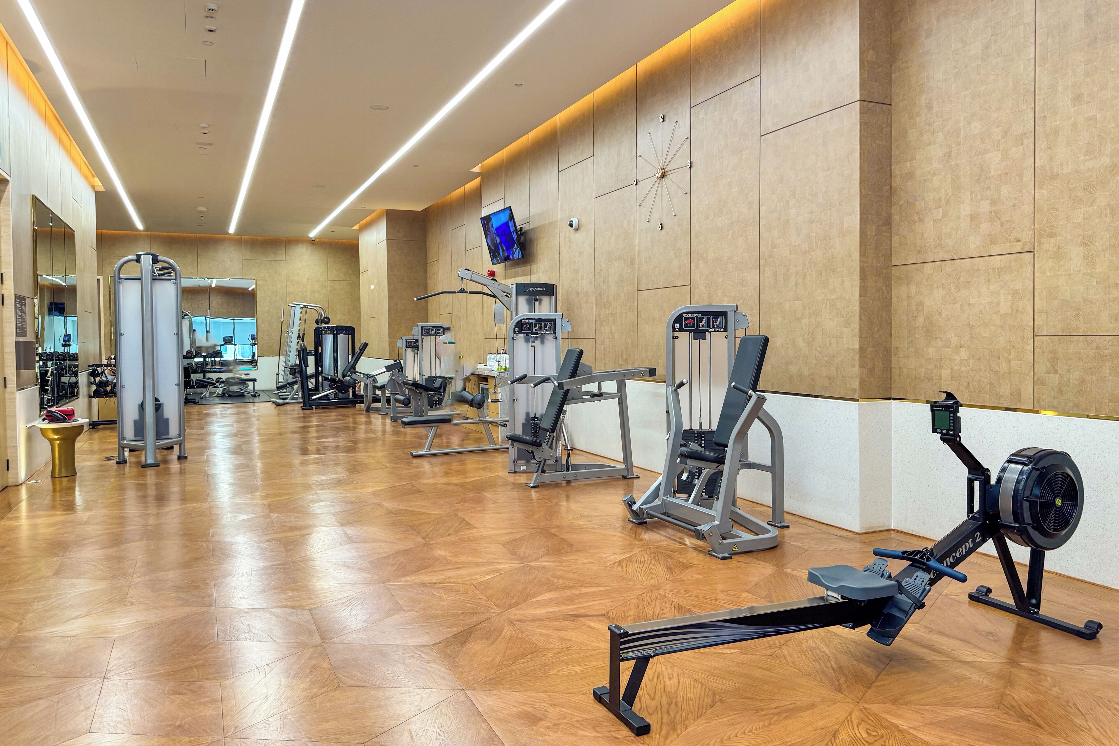A gym with various exercise machines, a TV mounted on the wall, and a wooden floor.