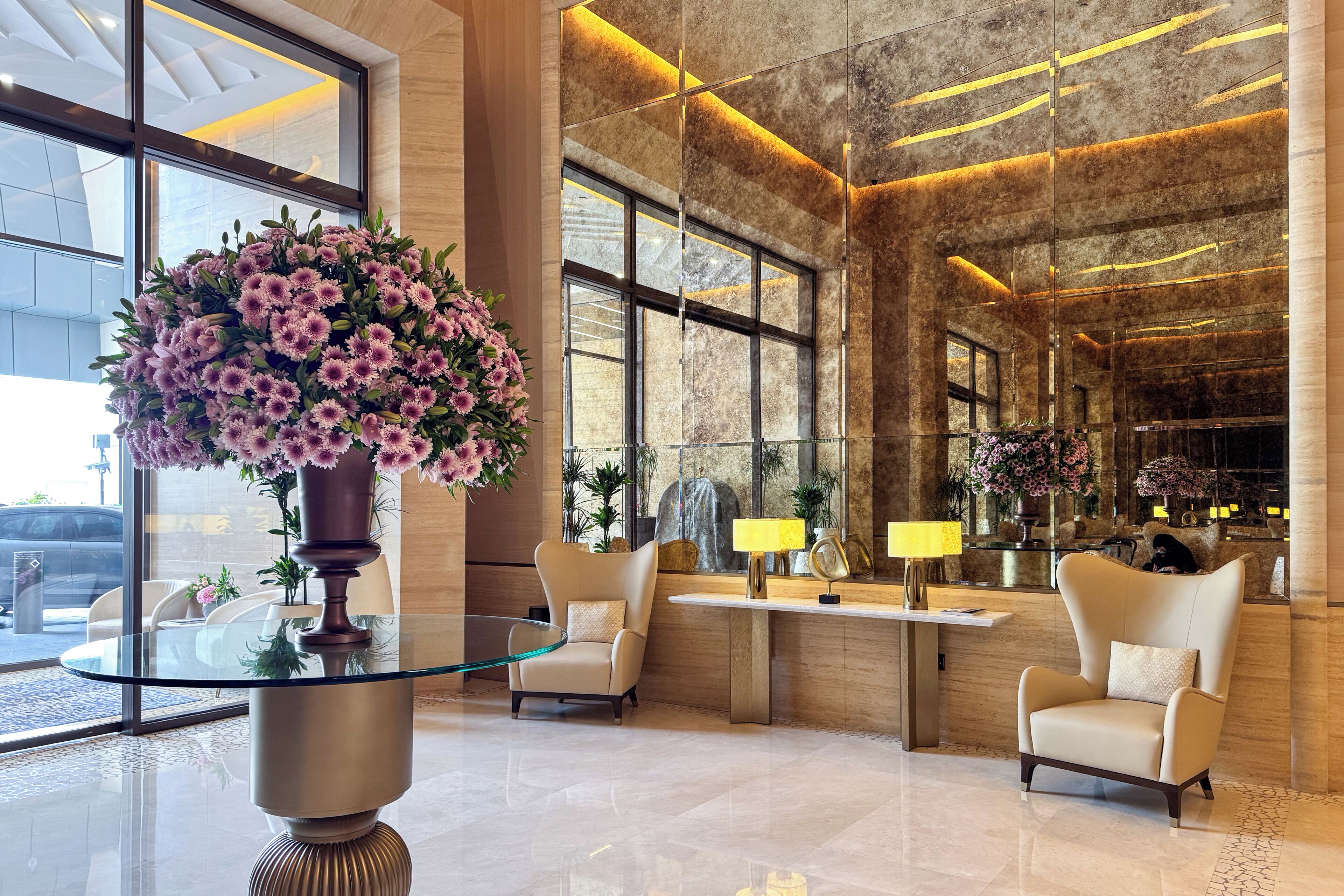 A modern lobby with a glass table, two armchairs, and a large floral arrangement.