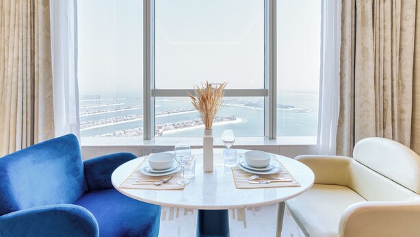 A dining area with a table set for two, a vase with dried plants, and a view of a coastal cityscape through large windows. - PVH - Modern Studio - Palm Tower (Dubai)