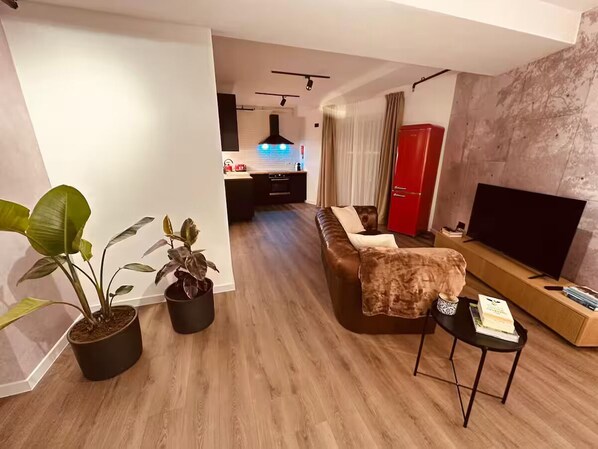 Una sala moderna con un sofá, una mesa de centro y un televisor. Se ven plantas en macetas y una cocina al fondo.
